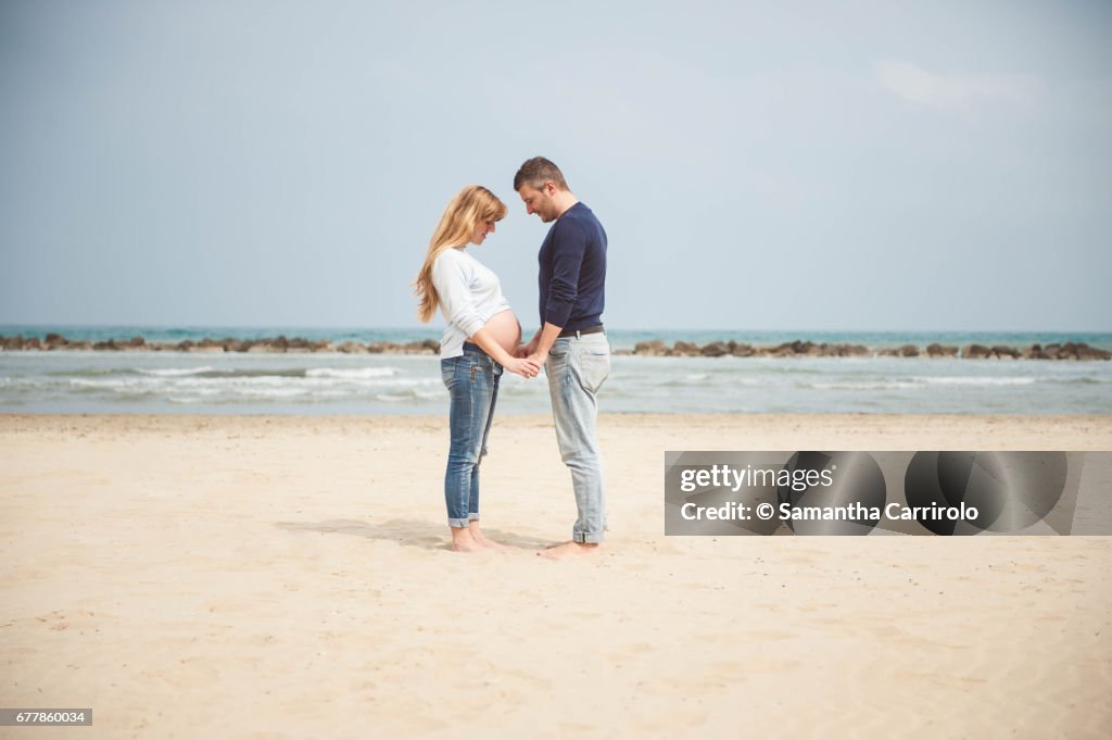Pregnant couple on the beach. Hand in hand. Looking at the belly. Casual clothes.