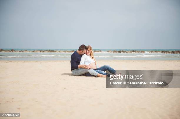 pregnant couple sitting on the beach. embrace. casual clothes. - coppia eterosessuale stock-fotos und bilder