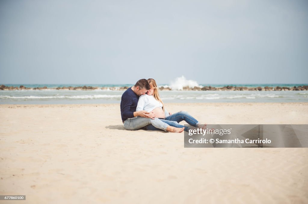 Pregnant couple sitting on the beach. Embrace. Casual clothes.