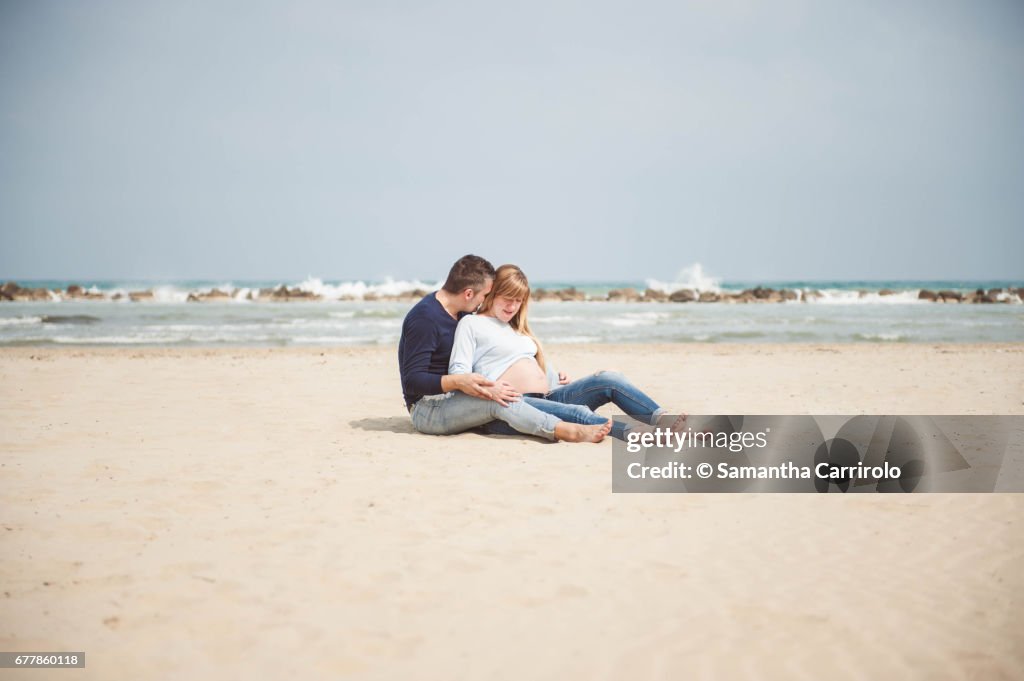Pregnant couple sitting on the beach. Embrace. Casual clothes.