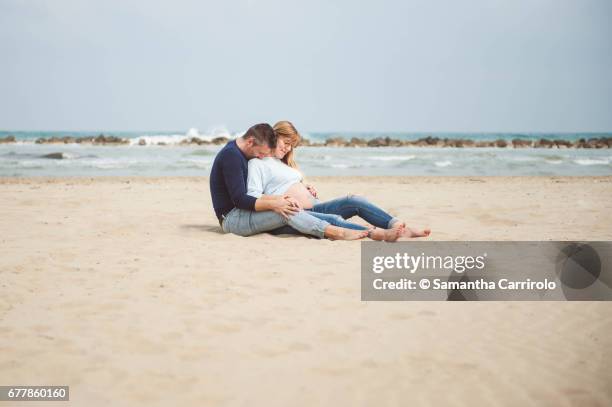 pregnant couple sitting on the beach. embrace. casual clothes. - serenità stockfoto's en -beelden