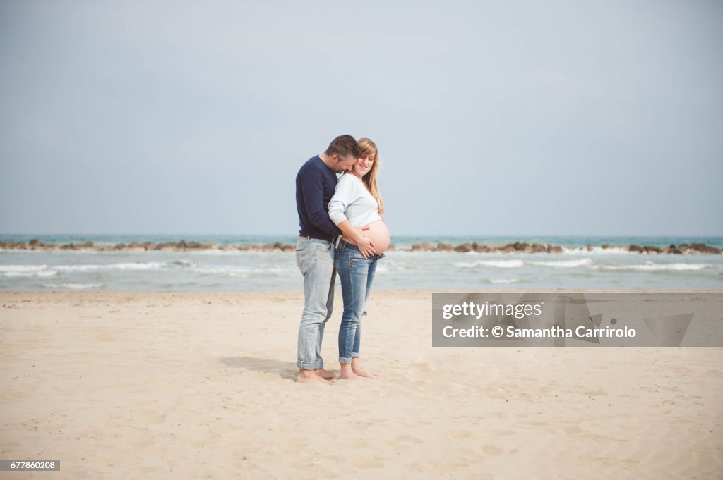 Pregnant couple on the beach. Hands on the belly. Embrace. Casual clothes.