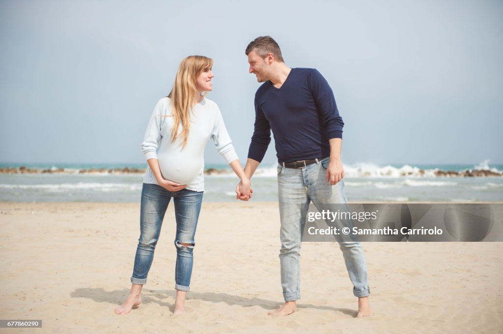 Pregnant couple on the beach. Hand in hand. Casual clothes. Hand on the belly.