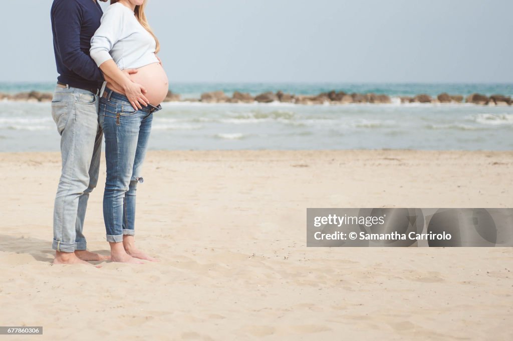 Pregnant couple on the beach. Hands on the belly. Embrace. Casual clothes.