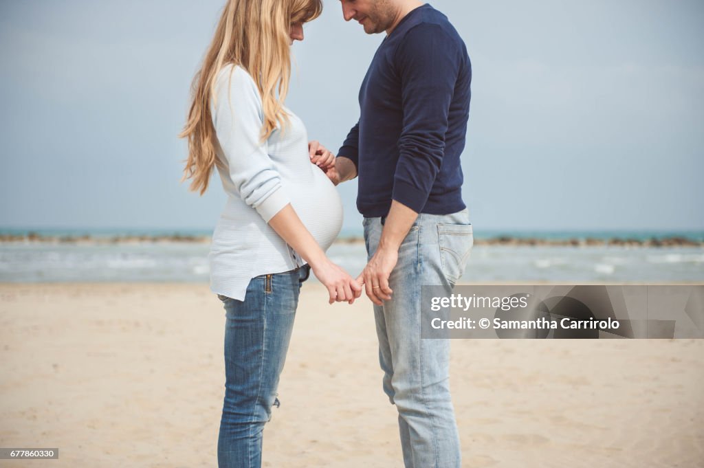 Pregnant couple on the beach. Hands on the belly. Casual clothes. Hand in hand.