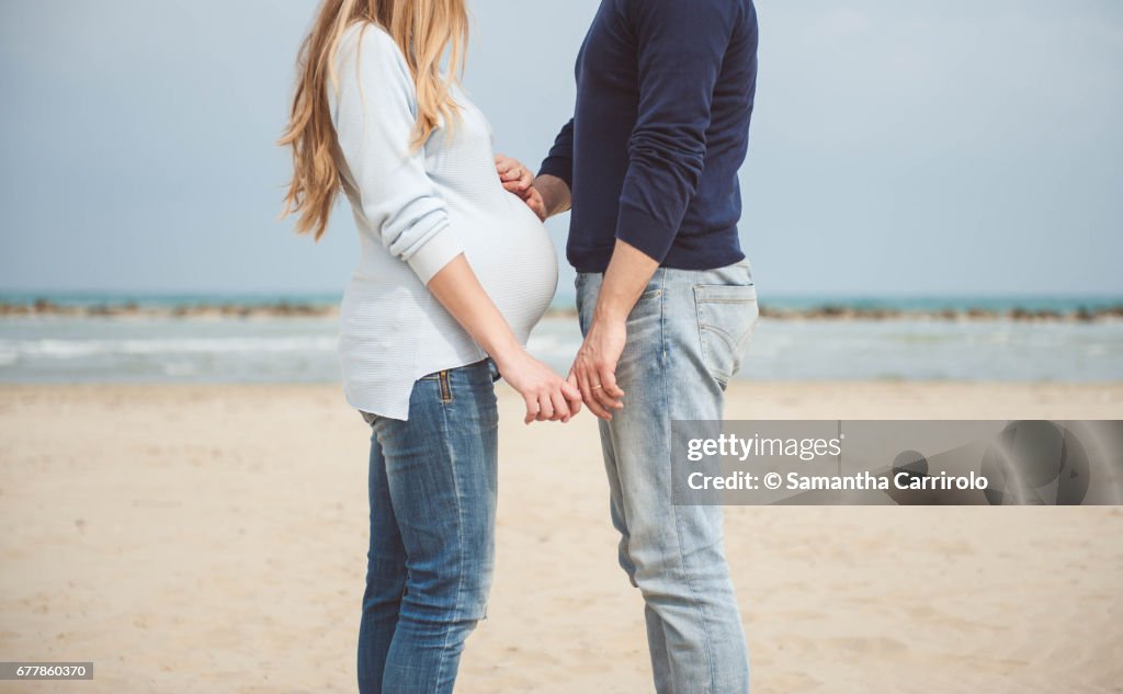Pregnant couple on the beach. Hands on the belly. Casual clothes. Hand in hand.