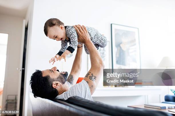 happy father lifting daughter while sitting on sofa at home - baby stockfoto's en -beelden