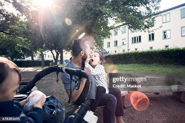 Father playing with daughter while sitting on bench by baby stroller at park