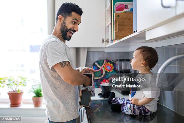 happy father making breakfast while looking at toddler sitting on kitchen counter - pai dono de casa - fotografias e filmes do acervo