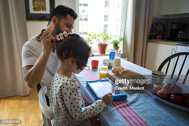 Girl using digital tablet while father tying her hair at table