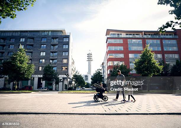 father walking with baby stroller looking at daughter riding push scooter in city - genderblend fotografías e imágenes de stock