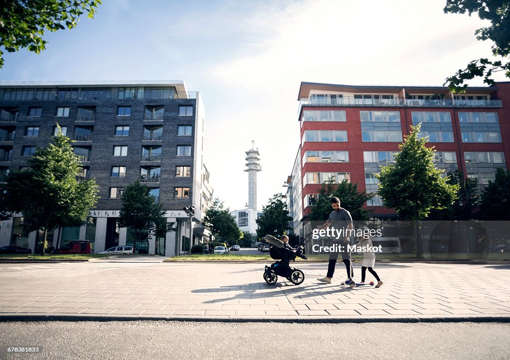 Father walking with baby stroller looking at daughter riding push scooter in city