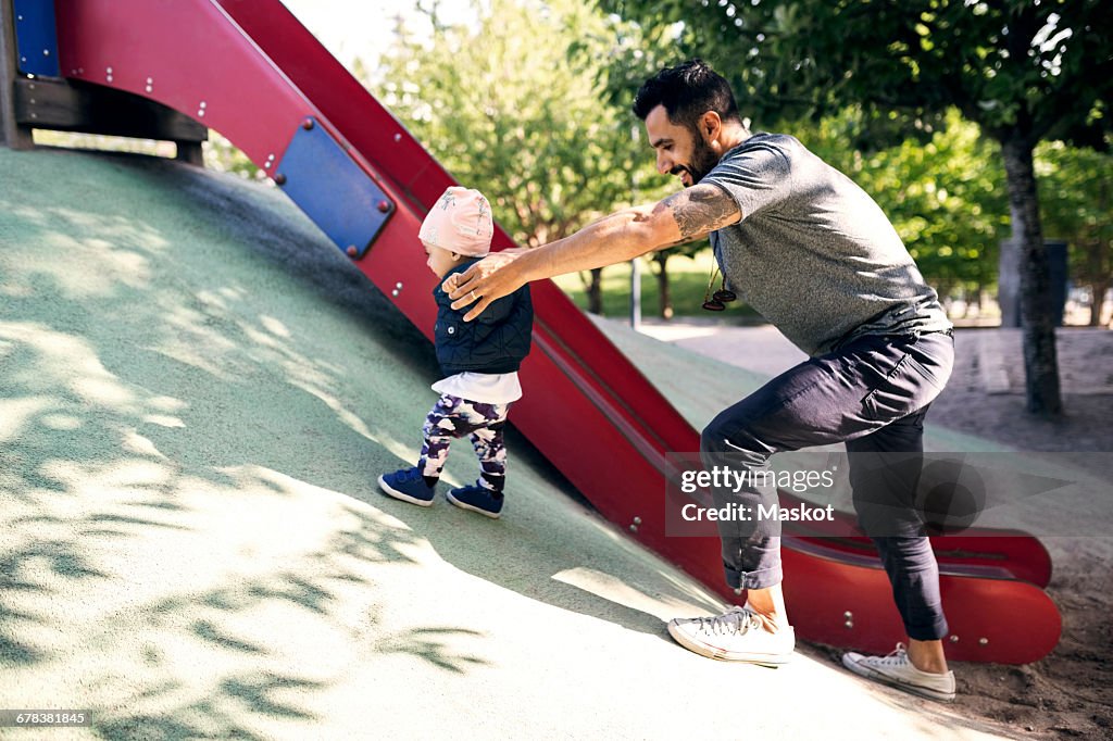 Happy father assisting daughter in climbing steep by slide at park