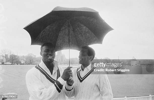 Barbadian cricketer Wes Hall and England cricketer Basil D'Oliveira share an umbrella during a hold-up in play between the West Indies and Worcester,...