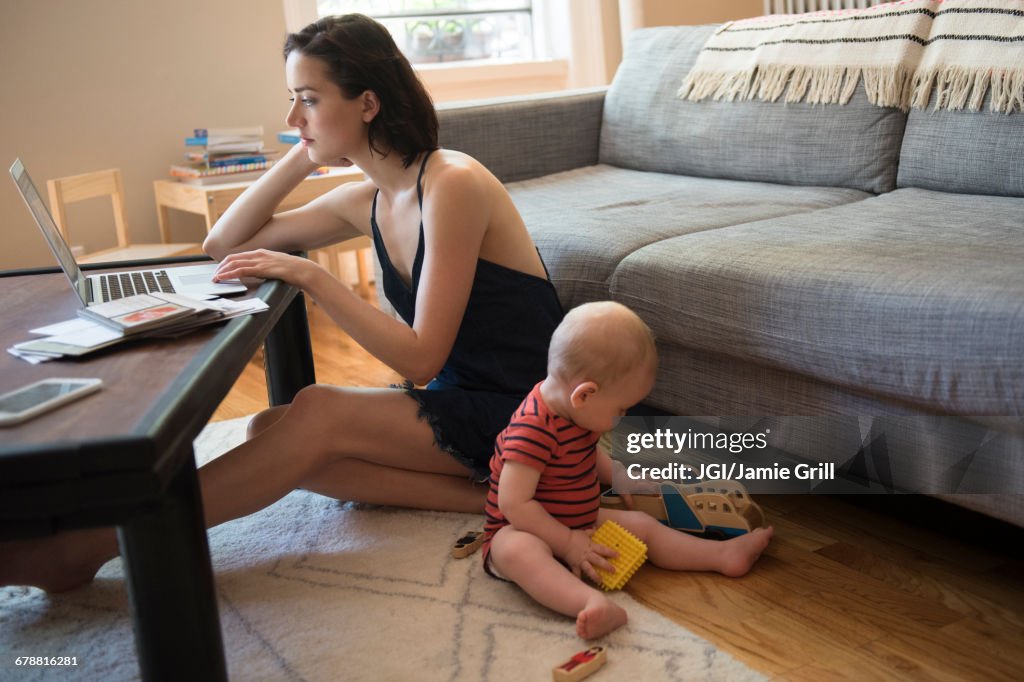 Caucasian mother sitting on floor using laptop near baby son