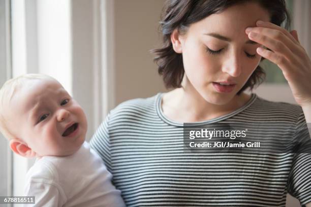 caucasian mother rubbing forehead while holding baby son - frustrated parent stock pictures, royalty-free photos & images