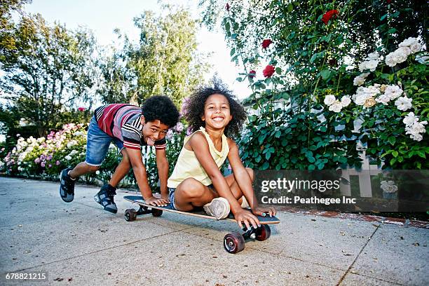 mixed race boy pushing sister on skateboard on sidewalk - summer press day ストックフォトと画像