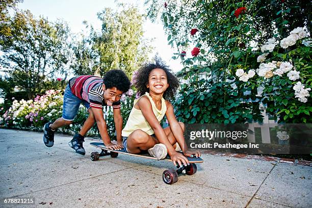 mixed race boy pushing sister on skateboard on sidewalk - boy skating stock pictures, royalty-free photos & images