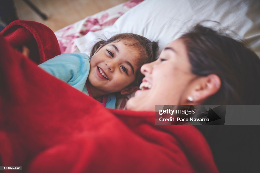 Laughing Hispanic mother and daughter under red blanket in bed