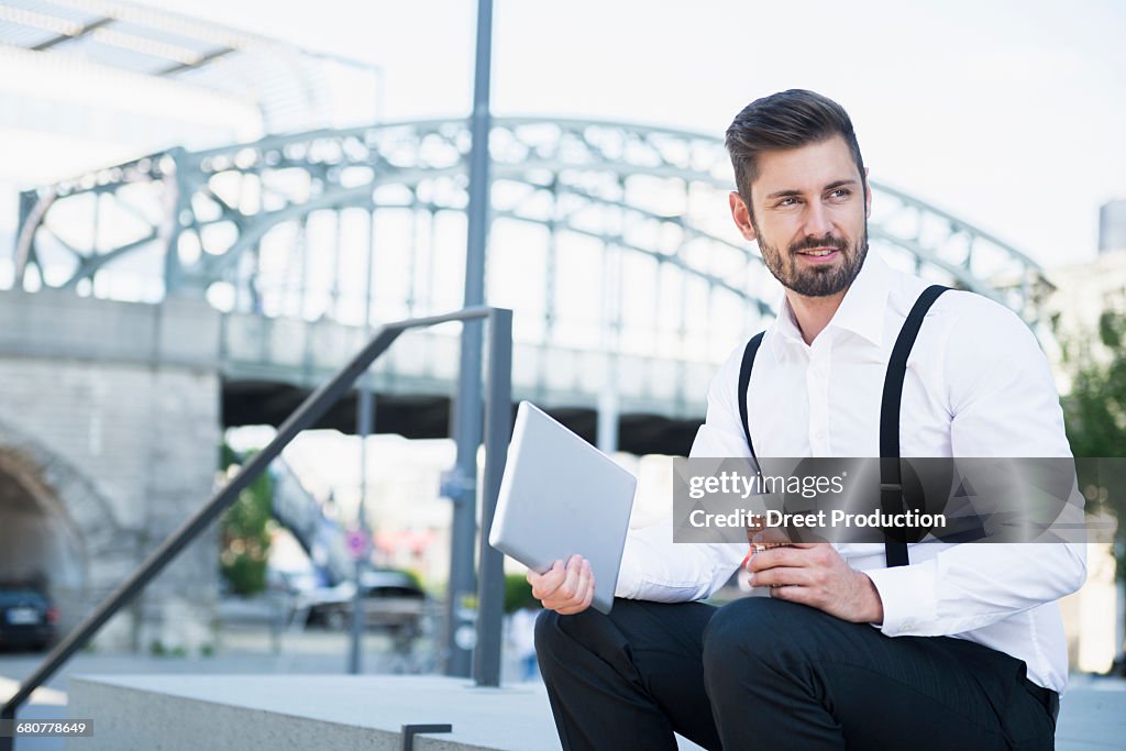 Businessman sitting on the stairs and holding digital tablet with coffee, Munich, Bavaria, Germany
