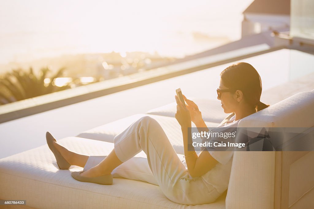 Woman using cell phone on chaise lounge at poolside on luxury patio