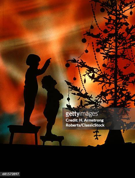 1920s 1930s TWO ANONYMOUS SILHOUETTED CHILDREN STANDING ON STOOLS LOOKING POINTING AT SKIMPY CHRISTMAS TREE