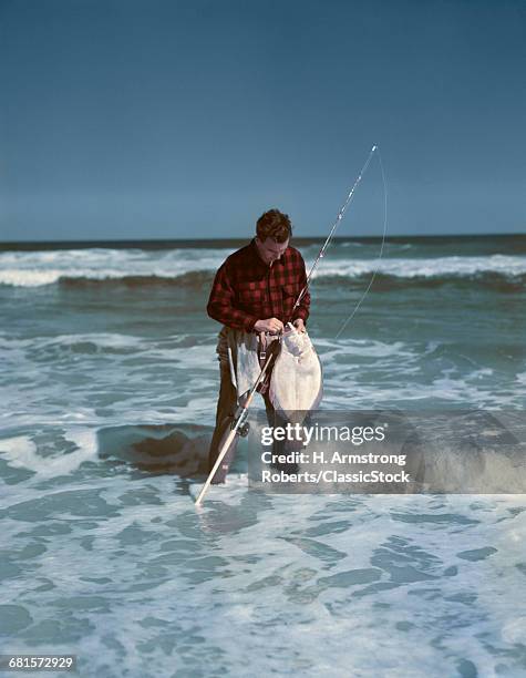 1940s 1950s MAN FISHING WEARING RED BLACK CHECKED SHIRT STANDING IN OCEAN SURF REMOVING FLOUNDER FROM HOOK