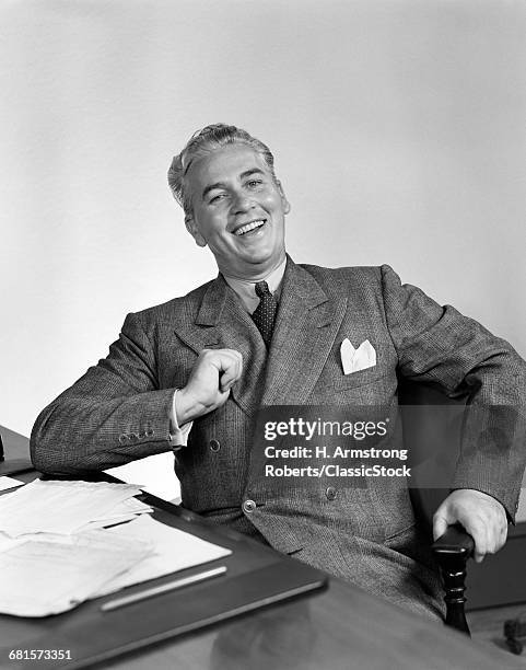 1940s HAPPY CONFIDENT BUSINESSMAN SITTING AT DESK THUMB TUCKED UNDER LAPEL IN GESTURE OF PRIDE LOOKING AT CAMERA