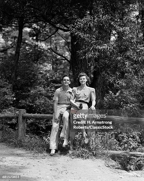 1930s 1940s TEENAGE COUPLE SITTING TOGETHER ON FENCE IN WOODS LOOKING AT CAMERA WOMAN HOLDING CAMERA