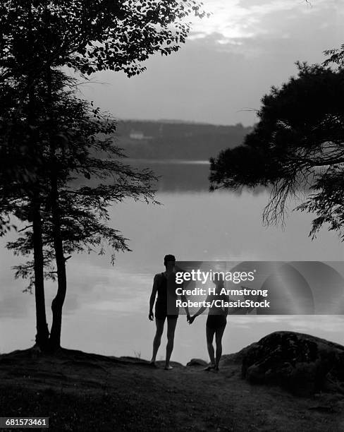 1930s REAR VIEW ANONYMOUS SILHOUETTED MAN AND WOMAN IN BATHING SUITS HOLDING HANDS WATCHING SUNSET LAKESIDE