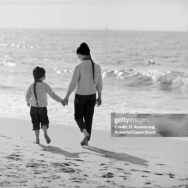1970s MOTHER DAUGHTER WALKING ALONG BEACH BACKS TO CAMERA HOLDING HANDS TALKING WEARING MATCHING JEANS SWEATERS CAPS BRAIDS