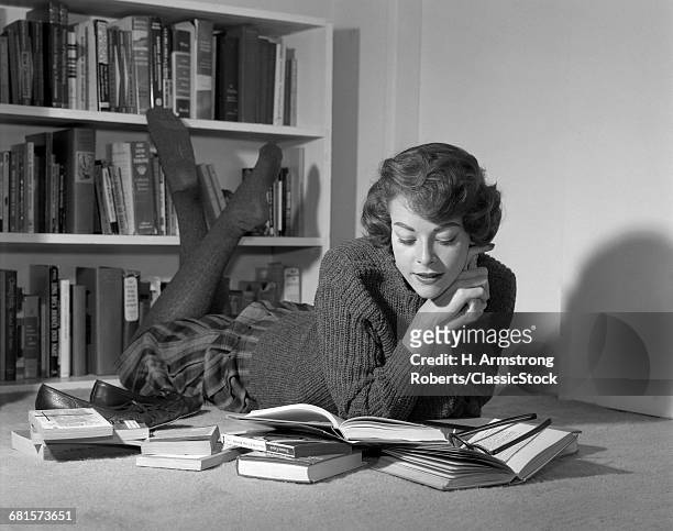 1960s YOUNG TEENAGE WOMAN LYING ON FLOOR IN FRONT OF BOOK SHELVES READING