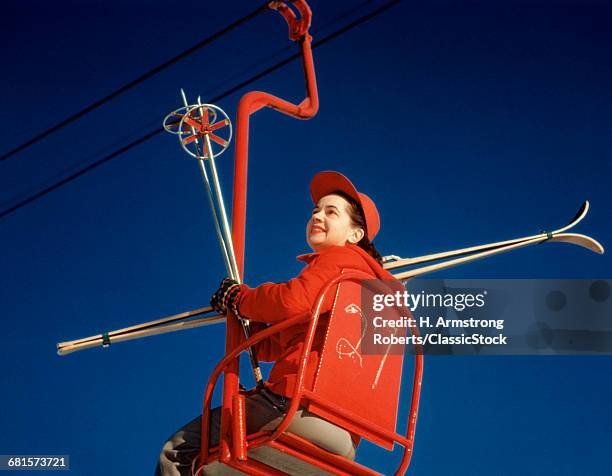 1950s BRUNETTE WOMAN RED SKI HAT JACKET SITTING IN RED SKI LIFT CHAIR HOLDING VINTAGE SKIS SKI POLES