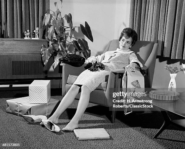 1960s WOMAN PLOPPED DOWN IN ARMCHAIR WITH SHOES KICKED OFF & PACKAGES AT FEET EXHAUSTED FROM SHOPPING