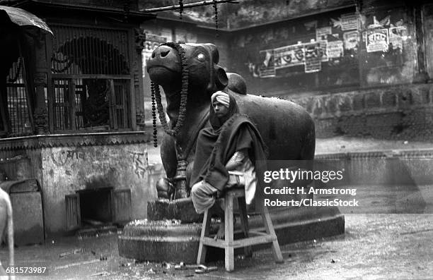 1920s 1930s BENARES INDIA MAN IN TURBAN SEATED BY STATUE OF NANDI SACRED HINDU GOD BULL RELIGION HINDUISM