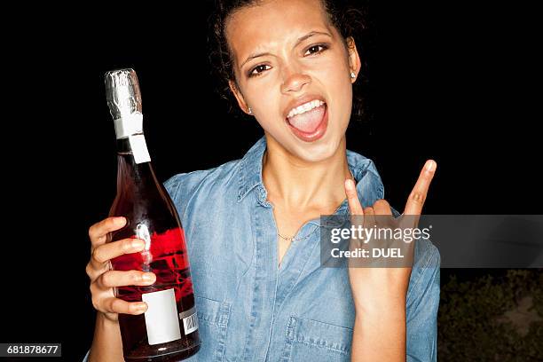 young woman holding bottle of wine doing rock sign looking at camera open mouthed - single rose stockfoto's en -beelden