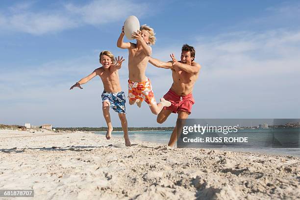 boy jumping with rugby ball chased by brother and father on beach, majorca, spain - familia con dos hijos fotografías e imágenes de stock