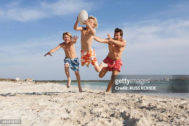 boy jumping with rugby ball chased by brother and father on beach, majorca, spain - tweens in bathing suits stock pictures, royalty-free photos & images