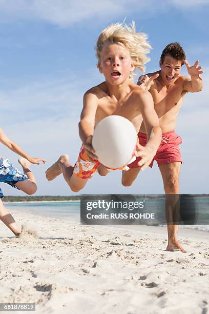 boy jumping mid air with rugby ball chased by brother and father on beach, majorca, spain - islas baleares fotografías e imágenes de stock