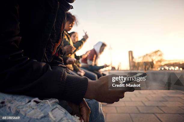 midsection of teenager using phone while sitting with friends by cobbled street - teenager smart phone foto e immagini stock