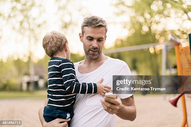 father using phone while carrying son at playground - father standing stockfoto's en -beelden