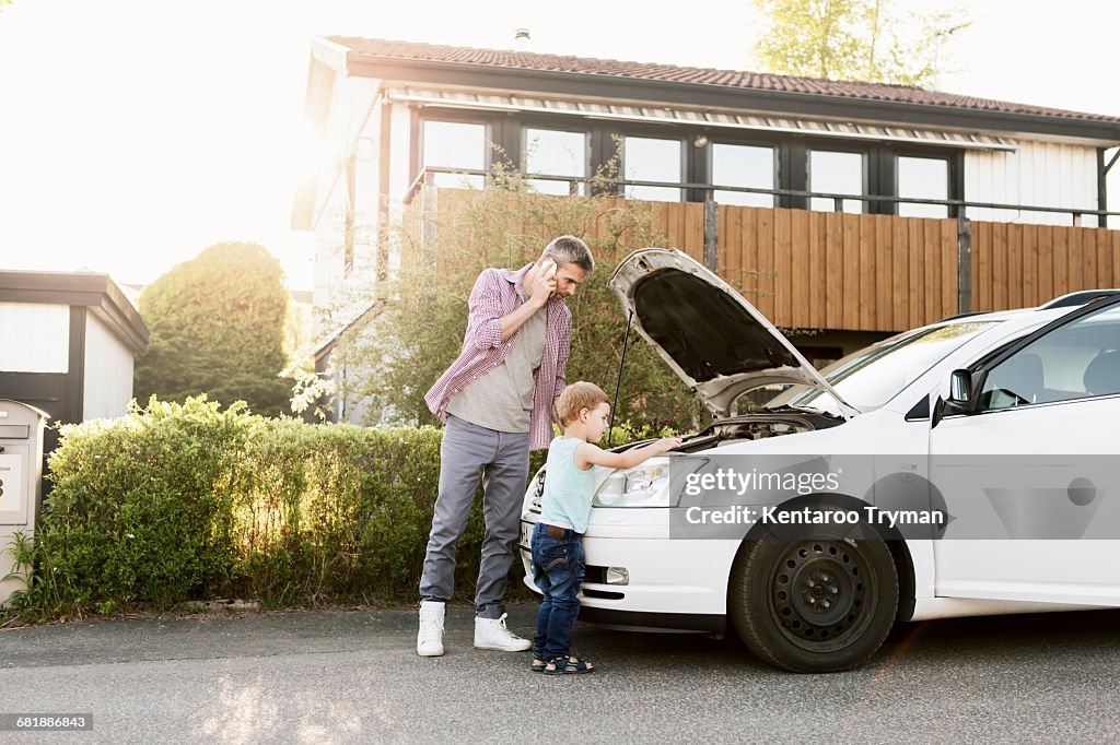 Father talking on phone standing with boy by broken down car on street