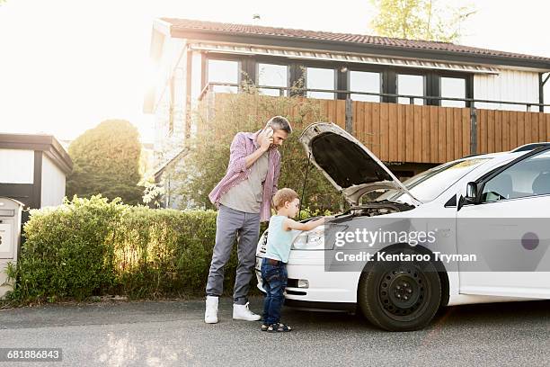father talking on phone standing with boy by broken down car on street - car broken down stock-fotos und bilder