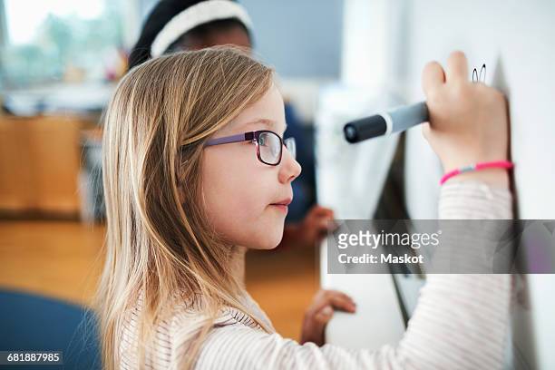 close-up of girl wearing eyeglasses writing on whiteboard in classroom - kids eyeglasses stock pictures, royalty-free photos & images
