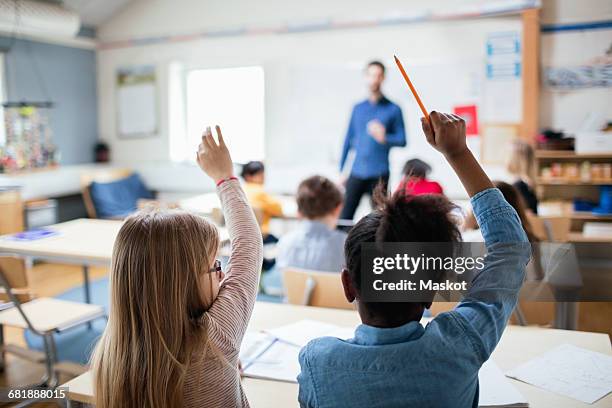 rear view of students sitting with hands raised in classroom - niveau primaire photos et images de collection