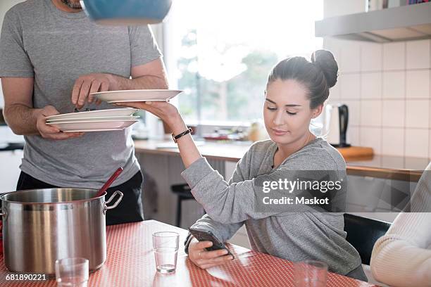 daughter giving plate to father while using smart phone at dining table - weekday stock-fotos und bilder