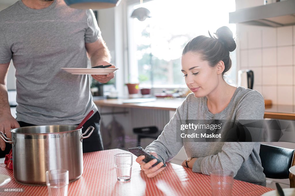 Midsection of father holding plate while daughter using smart phone at dining table in kitchen