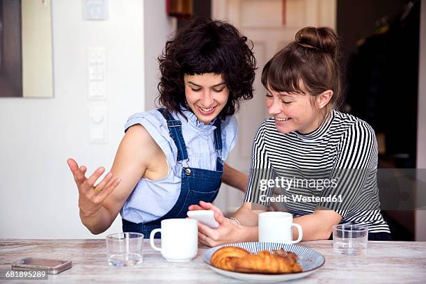 two women at breakfast table looking together at smartphone - woman kitchen stock-fotos und bilder