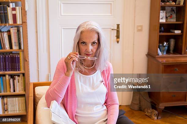 portrait of serious looking mature woman with newspaper sitting on couch at home - strictness stock pictures, royalty-free photos & images