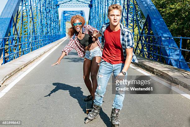 young couple with inline skates and skateboard riding on a bridge - patín en línea fotografías e imágenes de stock